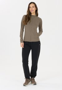 Beige Jacke mit durchgehendem Reißverschluss und langen Ärmeln, zwei Seitentaschen. Kombiniert mit schwarzen tapered Jogginghosen und braunen Schnürschuhen. Glatte Textur.