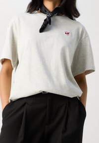 Jasnoszara bawełniana koszulka z krótkim rękawem i małym czerwonym haftowanym logo na piersi, zestawiona z czarnymi teksturowanymi spodniami w kroju dopasowanym.