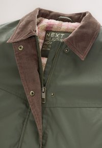 Zöld dzseki barna kordbársony gallérral, elülső cipzárral, pattintós gombokkal és rózsaszín kockás bolyhos béléssel, amely a nyaknál és a címkénél látható.