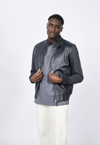 Chaqueta bomber de hombre, con frontal de cuero en gris oscuro, mangas de tela, puños acanalados, cremallera frontal, llevada sobre una camiseta gris y pantalones claros.