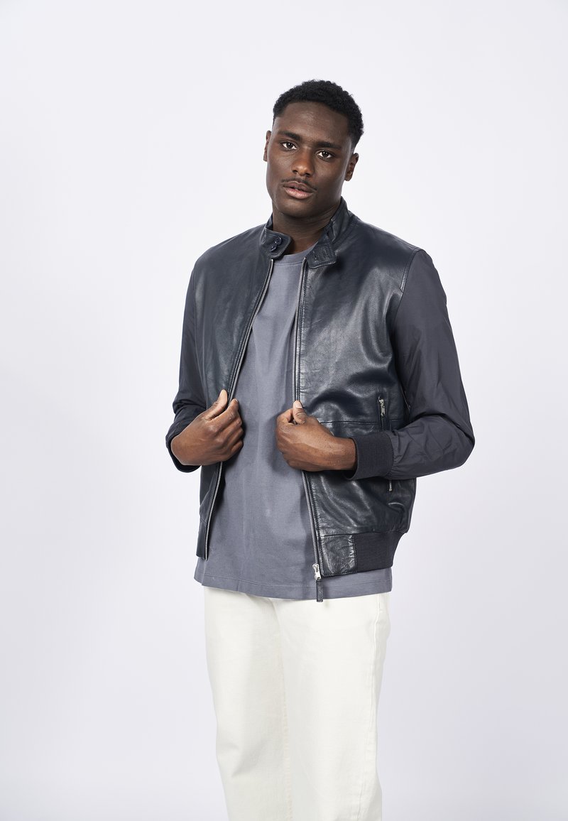 Chaqueta bomber de hombre, con frontal de cuero en gris oscuro, mangas de tela, puños acanalados, cremallera frontal, llevada sobre una camiseta gris y pantalones claros.