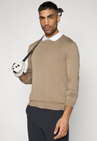 Μπεζ πλεκτή μπλούζα με στρογγυλό λαιμόκοψη, συνδυασμένη με λευκό πουκάμισο με γιακά. Το μοντέλο κρατά ένα μπαστούνι γκολφ φορώντας λευκό γάντι.