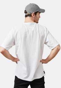 Camiseta de algodón blanca con mangas cortas, escote redondo y corte holgado. Un pequeño parche de logo oscuro es visible en la parte inferior izquierda.