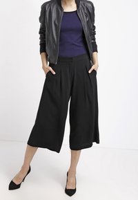 Schwarze, kurz geschnittene Culottes mit Falten, kombiniert mit einer dunklen Bomberjacke und einem marineblauen Oberteil. Das Outfit zeichnet sich durch glatte Stoffe und scharfe Silhouetten aus.