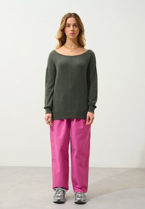 Pull vert foncé à encolure large et manches longues, associé à un pantalon rose ample et texturé, et des baskets grises.