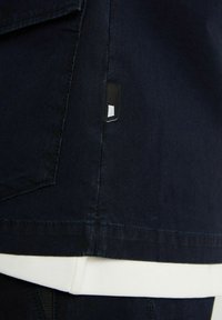 Navy blue jacket with visible stitching, featuring a black label on the side seam and a white inner lining visible at the hem.