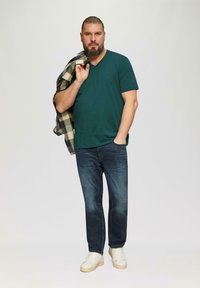 Grünes V-Ausschnitt-T-Shirt, dunkelblaue Jeans und weiße Sneaker. Ein graues und gelbes kariertes Hemd hängt über der Schulter. Einheitlicher Hintergrund.