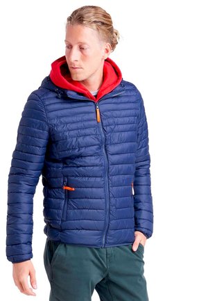 Un homme porte une veste matelassée bleu marine avec une capuche rouge et des détails de fermeture éclair orange sur fond blanc.