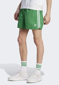 Green athletic shorts with white side stripes, made from lightweight fabric. Notable Adidas logo on the front. Worn with white sneakers and striped socks.