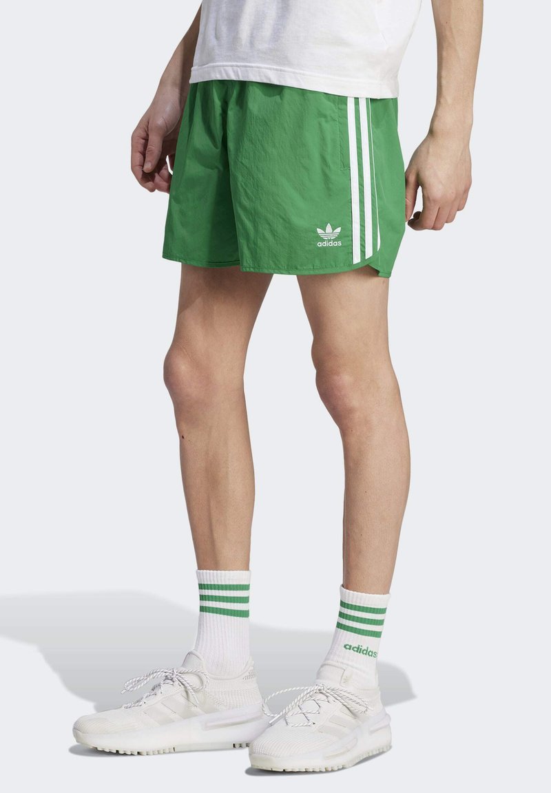 Green athletic shorts with white side stripes, made from lightweight fabric. Notable Adidas logo on the front. Worn with white sneakers and striped socks.