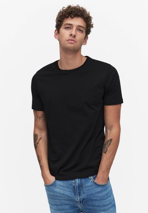 Młody mężczyzna o kręconych włosach, ubrany w prostą czarną koszulkę i niebieskie dżinsy, stojący z rękami w kieszeniach na białym tle.