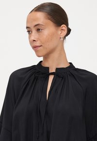Black blouse with a high collar, drawstring detail, and soft fabric. Features gathered shoulders and a relaxed silhouette. Silver earrings visible.