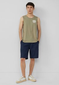 Olivgrünes ärmelloses Shirt mit "PACIFIC CUSTOMS" Grafik, kombiniert mit dunkelblauen Shorts. Dazu werden cremefarbene Sneakers mit beigen Akzenten getragen.