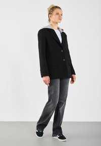 Čierny blazer s šedou kapucňou, jednoradové zapínanie, dva gombíky a bočné vrecká, v kombinácii s voľnými šedými džínsami a čiernymi teniskami.