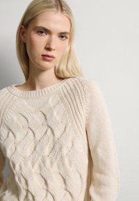 Suéter crema de punto trenzado con cuello redondo, patrones texturizados y acentos de canalé en los hombros y puños. Tejido suave y cálido.