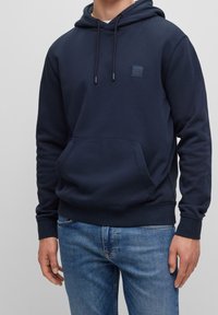 Navy blue hoodie in soft fabric with a kangaroo pocket, adjustable drawstrings, and a square logo patch on the chest. Paired with blue jeans.