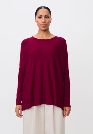 Frau steht, trägt ein lockeres, langärmeliges dunkelrotes Oberteil und weit geschnittene weiße Hosen, vor einem schlichten hellen Hintergrund.