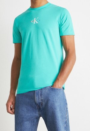 T-shirt di cotone turchese con un piccolo logo bianco sul petto, maniche corte e scollo rotondo classico, abbinato a jeans azzurri chiari.