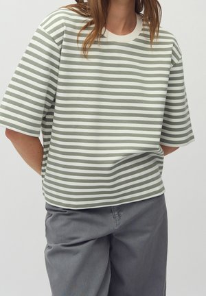 Persoon die een los, wit T-shirt met korte mouwen met grijze horizontale strepen en een grijze broek draagt, staand met de handen achter de rug.