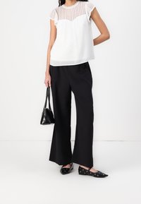 Žena v bílém krajkovém topu, černých širokých kalhotách a černých špicatých balerínách s kovovými očky, držící malou černou kabelku.