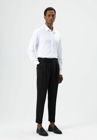 Chemise blanche à manches longues avec col, associée à un pantalon noir taille haute avec détails de boutons sur le côté et revers retroussés. Des mocassins noirs complètent la tenue.