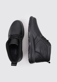 Un par de botas de cuero negro con cordones hasta el tobillo, una mostrando la vista superior y la otra el perfil lateral, sobre un fondo claro liso.