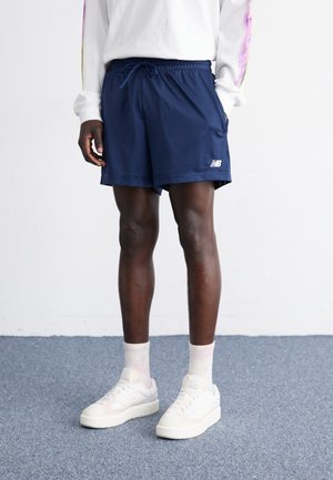 Mann trägt marineblaue, sportliche Netzshorts, weiße Socken, weiße Turnschuhe und ein weißes Langarmshirt mit bunten Ärmeln und steht auf einem blauen Teppich.