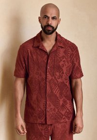 Chemise à manches courtes texturée rouge foncé avec motifs géométriques, motifs de palmiers et fermeture boutonnée ; dotée d'une poche poitrine unique.