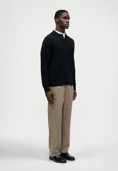 Camisa polo preta de manga longa, tecido de malha, punhos canelados. Calças texturizadas bege, corte solto. Sapatos pretos, superfície lisa. Fundo neutro.