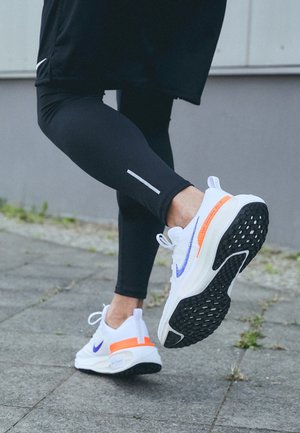 Białe buty sportowe z fioletowymi i pomarańczowymi akcentami, z teksturowaną czarną podeszwą. Noszone z czarnymi legginsami, widocznymi z odblaskowymi detalami.