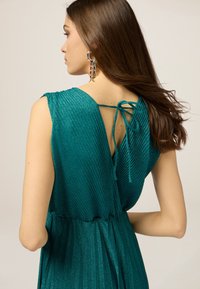 Türkisfarbenes, ärmelloses Kleid mit einem strukturierten Rippenmuster, das einen tiefen V-Ausschnitt auf der Rückseite und eine gebundene Detail aufweist. Glänzendes, leichtes Material.
