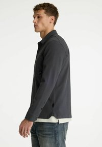 Dunkelgraues Herrenhemd zum Knöpfen mit strukturiertem Muster, leichtem Glanz, klassischem Kragen und langen Ärmeln. Verfügt über eine Brusttasche und einen abgerundeten Saum.