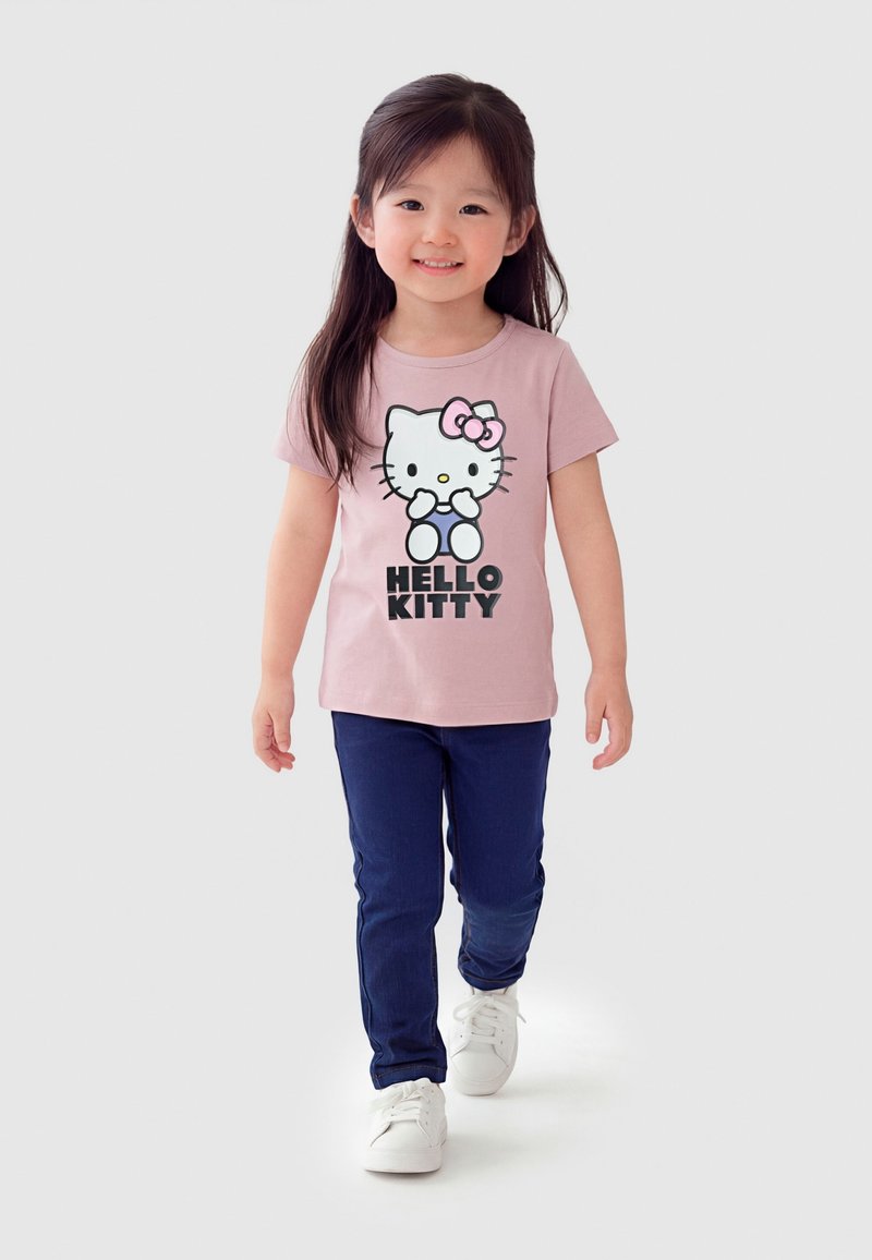 Mladé dievča kráča, oblečené v ružovom tričku Hello Kitty, tmavomodrých nohaviciach a bielych teniskách, na jednoduchom svetlosivom pozadí.