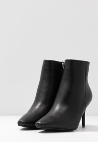 Schwarze Leder-Ankle-Boots mit spitzem Zehenbereich, schlanken hohen Absätzen und seitlichen Reißverschlüssen, auf einer weißen Oberfläche vor einem schlichten Hintergrund präsentiert.