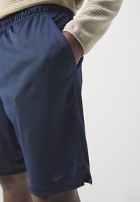 Närbild av en person som bär marinblåa träningsshorts med Nike-logotyp, handen vilande i sidofickan, och en beige ribbad långärmad tröja.