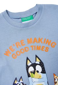 Jasnoniebieska koszula z napisem „Tworzymy dobre chwile” w kolorze pomarańczowym nad trzema kreskówkowymi psami, z których jeden trzyma zielony liść, zabawny wzór.