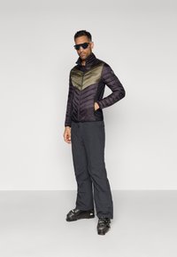 Piepildīta jaka ar melnu augšdaļu un olīvzaļās krāsas chevron dizainu, apvienota ar melniem slēpošanas biksēm un melniem slēpošanas zābakiem. Tiek iekļautas saulesbrilles.