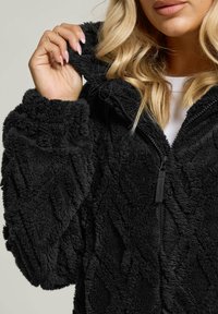 Musta fleece-tyylinen vetoketjullinen takki, jossa on pehmeä, teksturoitu pinta, huppu ja vetoketjusulku, esittelee hienovaraisia geometrisia kuvioita.