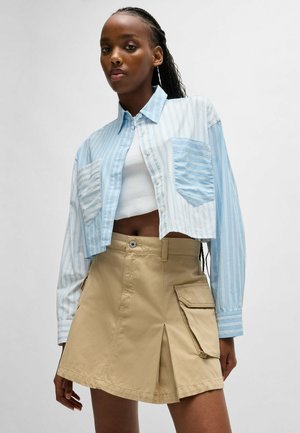 Chemise courte à rayures bleu clair et blanches avec deux poches avant ; associée à une jupe trapèze beige avec poches latérales et plis.