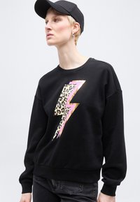 Άτομο που φοράει μαύρο φούτερ με λεοπάρ σχέδιο και ροζ αστραπή, μαύρα τζιν, μαύρο καπέλο και ασημένια σκουλαρίκια κρίκους.