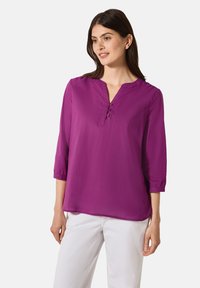 Lila Bluse mit Dreiviertelärmel, V-Ausschnitt und Knopfdetails. Glattes, leichtes Material. Kombiniert mit weißen Hosen.
