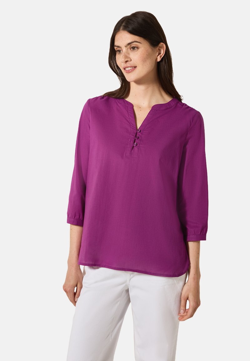 Lila Bluse mit Dreiviertelärmel, V-Ausschnitt und Knopfdetails. Glattes, leichtes Material. Kombiniert mit weißen Hosen.