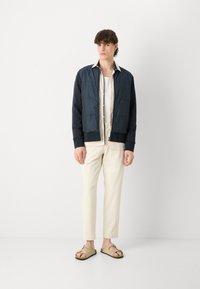 Chaqueta de plumas azul marino con textura suave, cuello alto y bolsillos frontales, combinada con una camisa ligera y pantalones cremosos. Sandalias suaves.