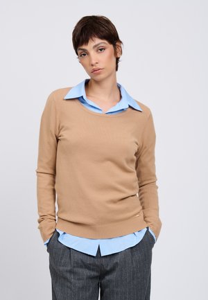 Junge Person mit kurzen braunen Haaren trägt einen beigen Pullover über einem hellblauen Hemd mit Kragen und dunkle, fein gestreifte Hose, Hände in den Taschen.