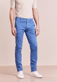 Mäns blå bomullbyxor med slim fit, med knäppning fram med knapp och dragkedja, bälteshällor och sidofickor; i kombination med vita sneakers.