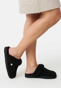 Sorte slip-on tøfler lavet af ruskind med en fluffy pelskrave. Tekstureret sål for godt greb. Simpelt design, ingen synlige mønstre eller accenter.