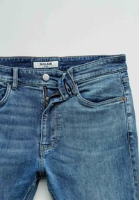 Blauwe skinny jeans met gedeeltelijk opengemaakte ritssluiting, knoop vast, en zichtbare voorzakken op een lichte achtergrond.