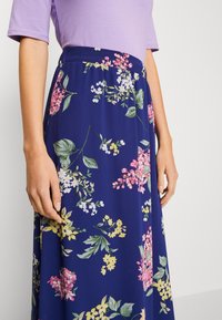 Kvinna klädd i en lila topp och en marinblå kjol med rosa, gula och vita blommönster, stående mot en ljus, enfärgad bakgrund.