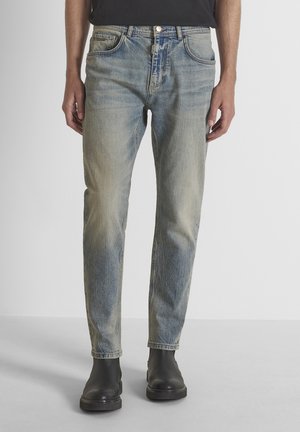 Persona con vaqueros azules descoloridos de corte relajado y botines negros de tobillo, de pie frente a un fondo sencillo.