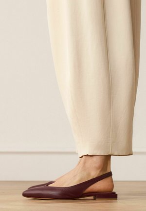 Vrouw draagt beige cropped broek en donkerpaarse slingback ballerina’s, staand op een houten vloer tegen een effen muur.